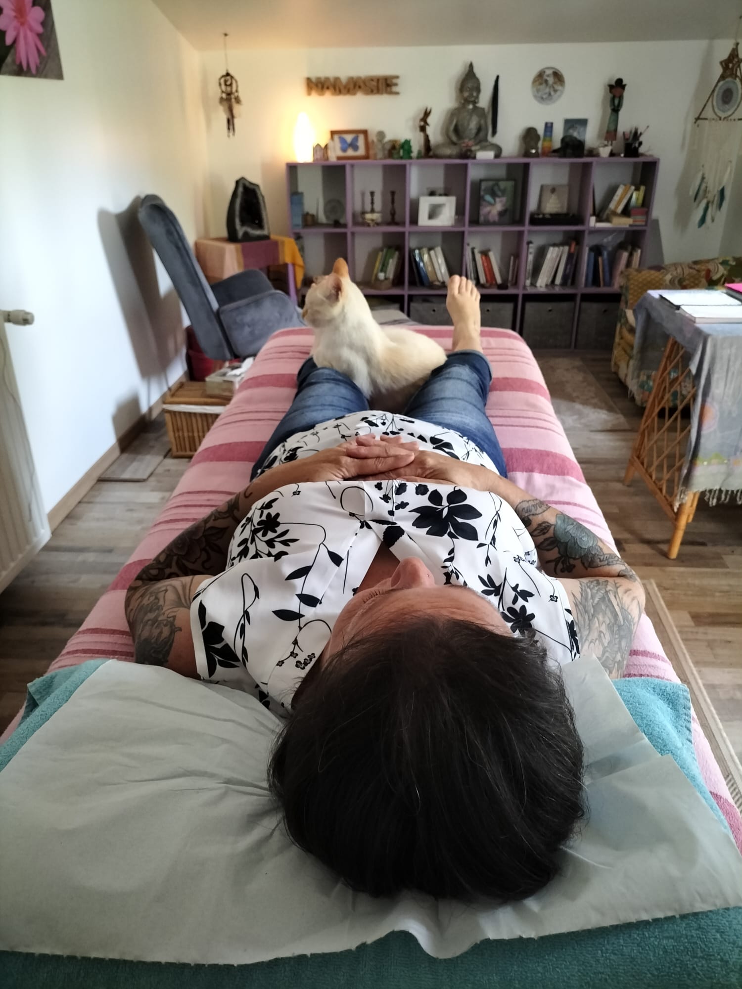 Personne allongée sur une table de soin, tête tournée vers le drap blanc, pieds nus sur le sol. Ambiance calme et zen dans une pièce où l’on aperçoit des étagères remplies de livres et objets spirituels, un chat blanc allongé près des pieds et des éléments décoratifs sur les murs.