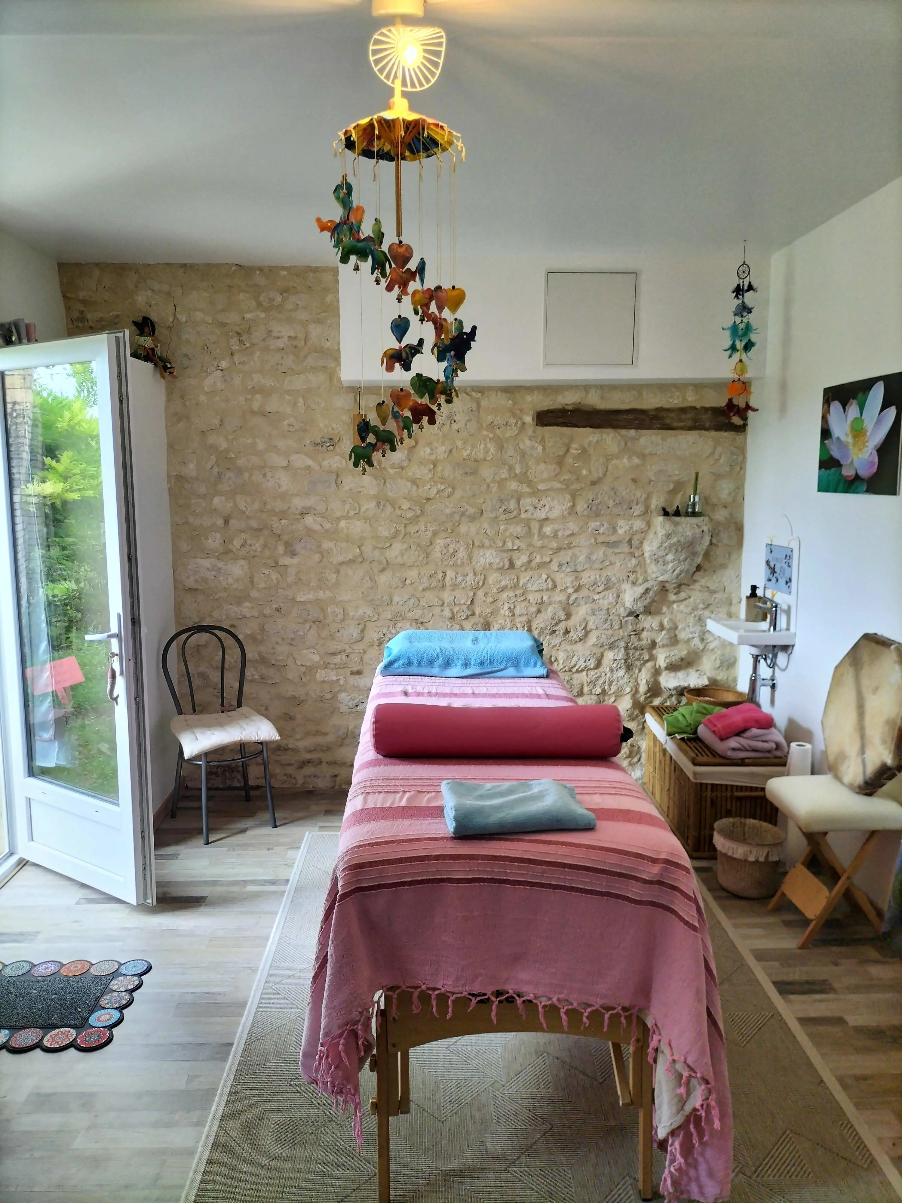 Salle de soins chaleureuse et lumineuse avec table de soin, couvertures colorées et coussins, mur en pierres, ouverture sur l’extérieur.