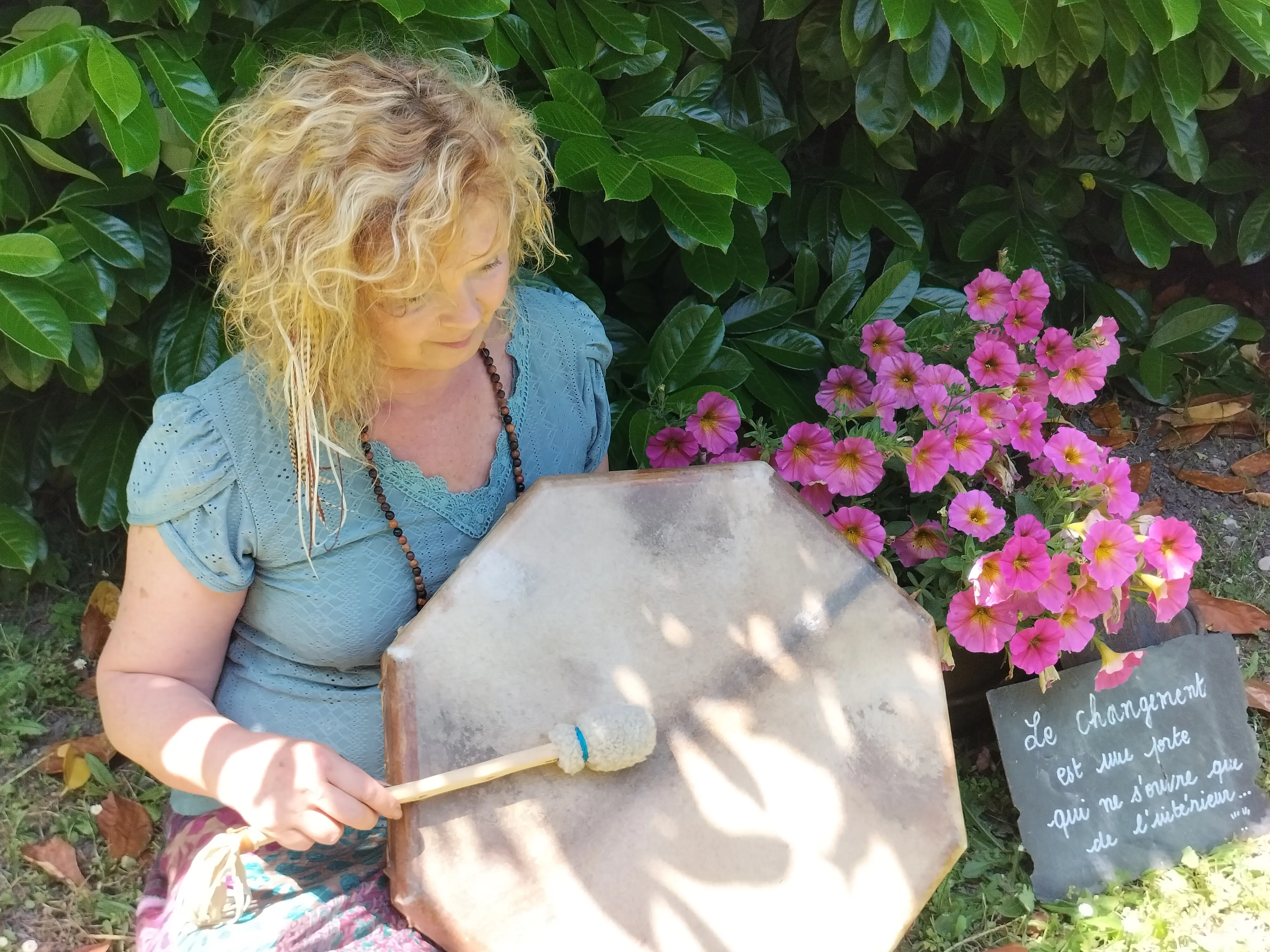 Sophie Latour médium en plein air, tenant un grand gong en métal dans un jardin, entourée de fleurs roses et de feuillages verts.