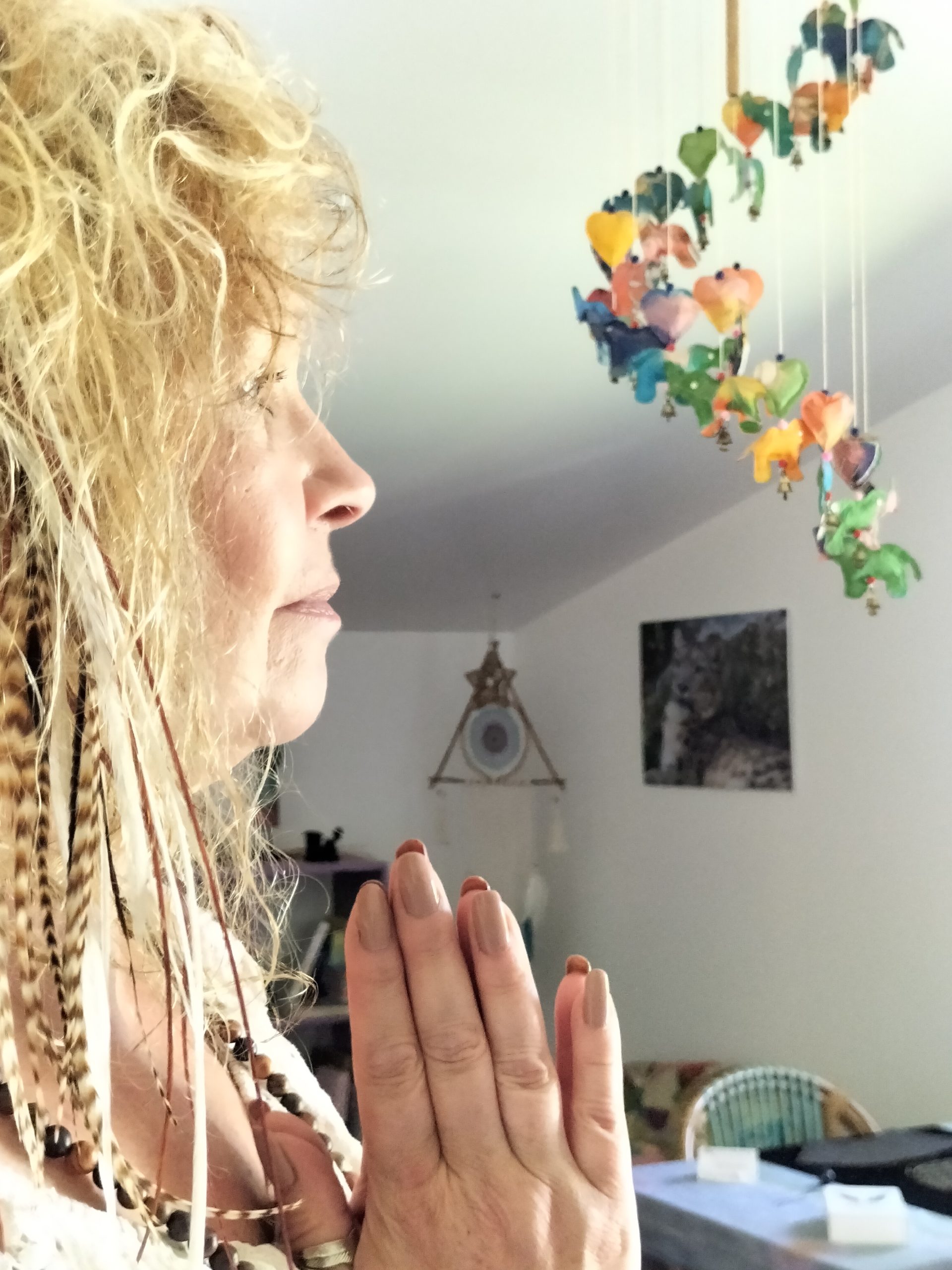 Une femme priant avec les mains jointes, regard tourné vers un mobile pendule coloré suspendu dans une pièce lumineuse. Intégrer l’idée d’initiation pendule radiesthésie.