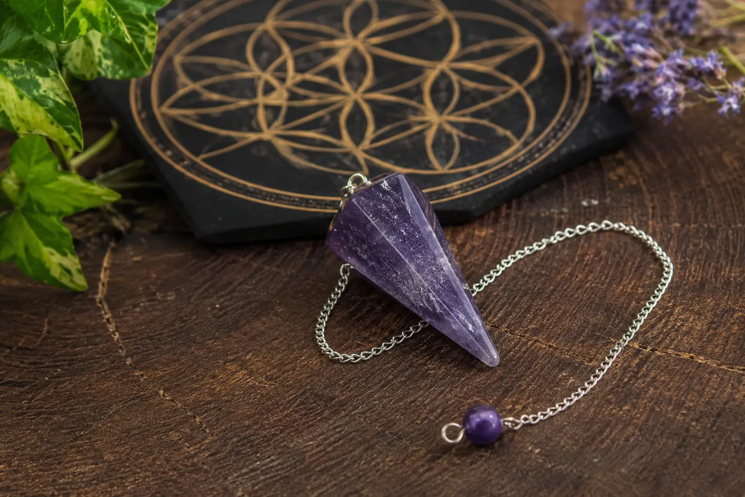 Pendentif en améthyste violet en forme de pointe sur chaîne argentée, posé sur une surface en bois avec un tapis ésotérique géométrique et des plantes.