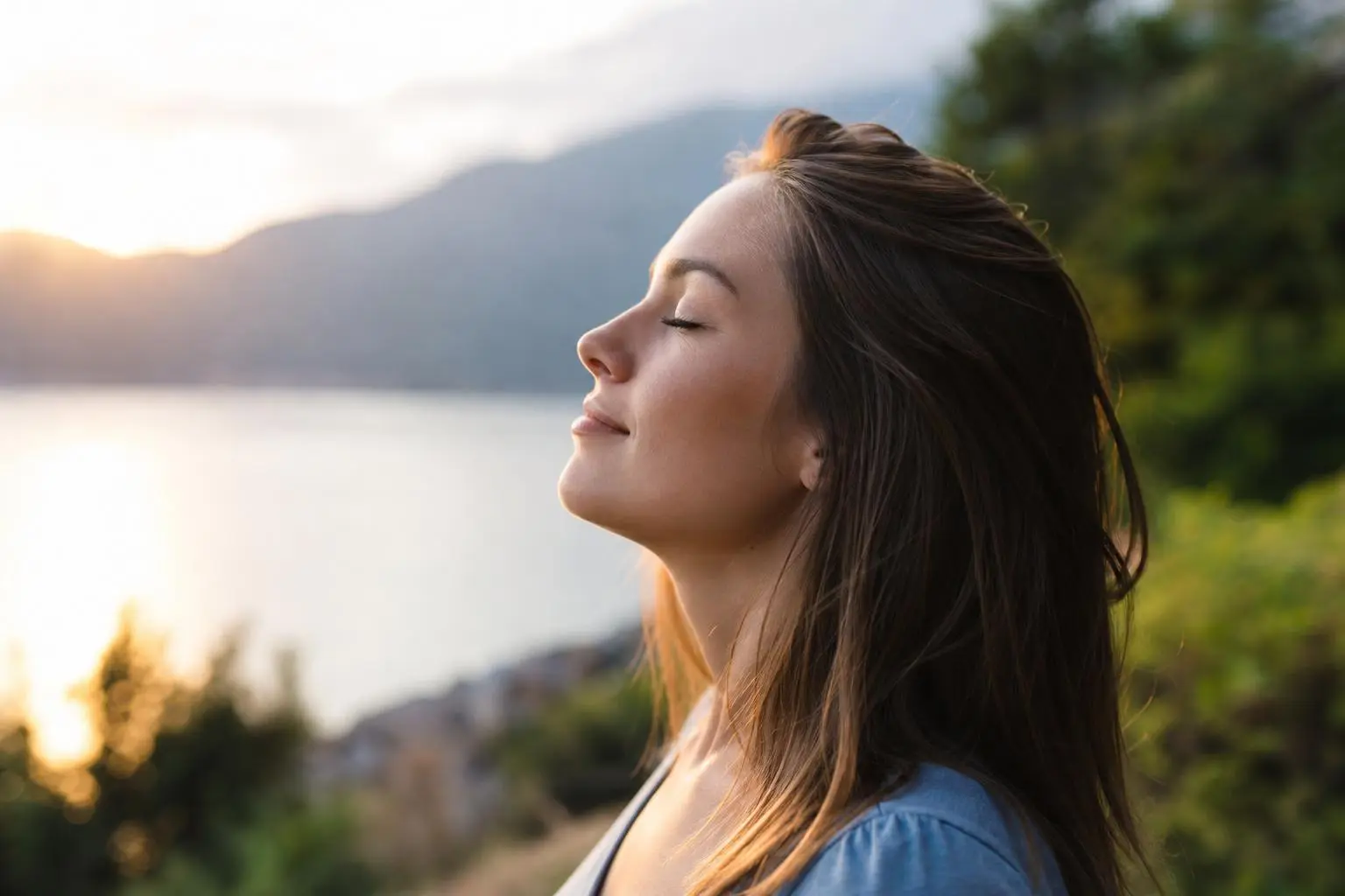 Poignée d’un visage souriant, femme en profil, yeux fermés, respirant calmement au lever ou coucher de soleil sur un paysage lacustre et montagneux. intègre comment retrouver son équilibre émotionnel.