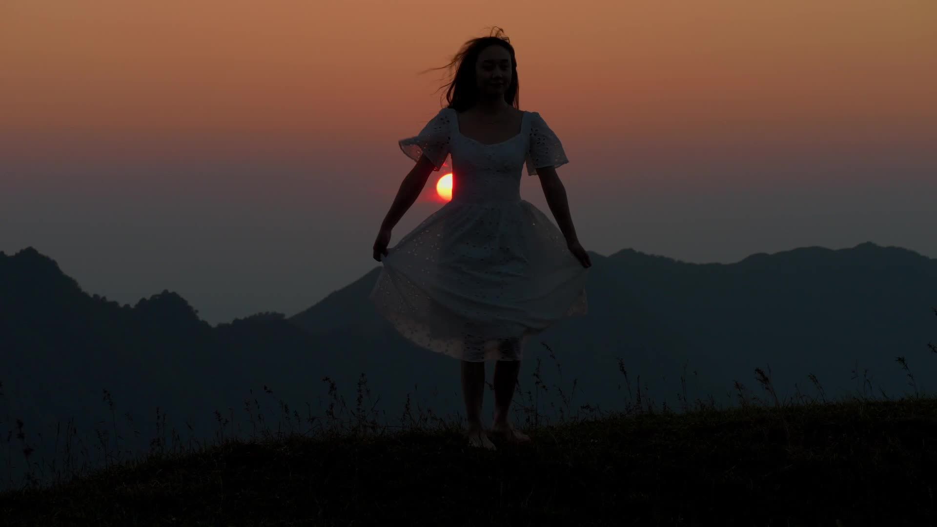 Une silhouette de femme en robe tourne sur une colline au coucher du soleil, avec le soleil orange en arrière-plan.