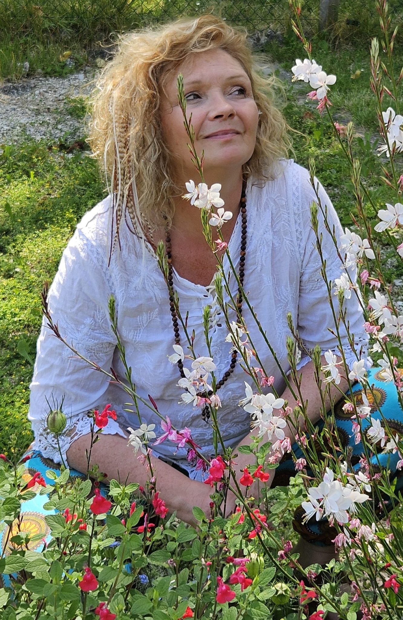 Une femme assise dans un jardin parmi des fleurs, regardant vers le haut avec un sourire.
