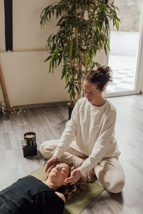 Une thérapeute réalise une séance de soin énergétique sur une cliente allongée sur un tapis de yoga dans une pièce lumineuse.
